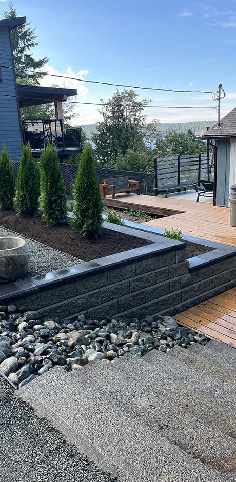 Modern landscaped patio featuring shrubs, stones, wooden deck, and scenic waterfront view.