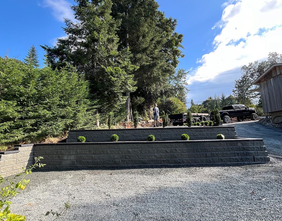 Landscaped yard with stone retaining walls, greenery, and a worker in the background.